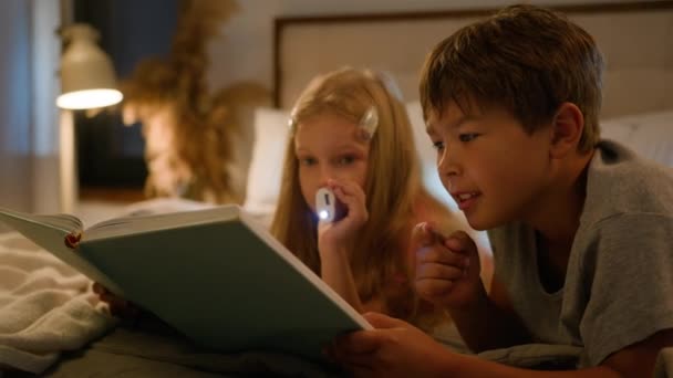 Caucasian children kids boy girl older brother younger sister lying on bed at home evening reading interesting book using flashlight read fairytale story for night bedtime happy family relationships - Materiał filmowy, wideo