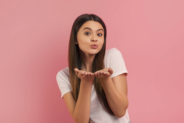 Charming teen girl blowing a playful kiss at camera, hands extended forward, beautiful female teenager with affectionate expression standing against pink background, symbolizing love and kindness - Fotografie, Obrázek