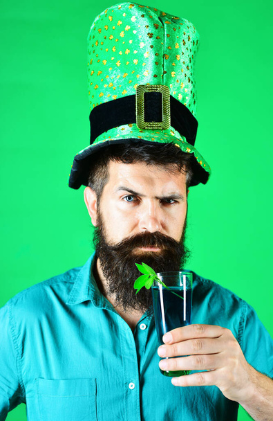 Saint Patricks day celebration. Bearded man in leprechaun hat with glass of green beer. Patrick day pub party. Closeup portrait serious man in green top hat drinking beer. Glass of beer with shamrock - Фото, изображение