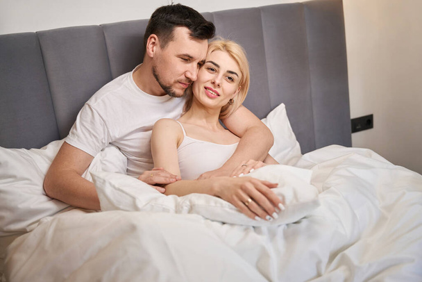 Young female is basking in the arms of her lover, the couple is seated on a comfortable bed - Foto, Imagem