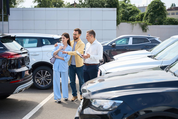 Husband and wife choose a car in the yard of a car dealership, a consultant helps them - Fotó, kép