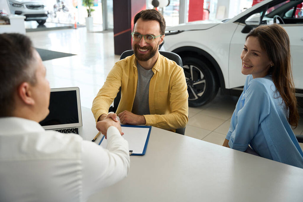 Couple communicates with a consultant in the office area of a car dealership, the men greet each other - Foto, Bild