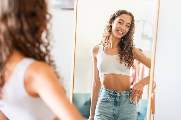 Weight Loss Concept. Happy Young Teen Age Girl Wearing Jeans After Dieting, Comparing Size Before And After Slimming, Posing Near Mirror Standing At Home. Selective Focus, Copy Space - Фото, изображение
