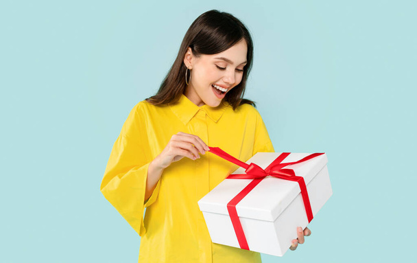 Excited emotional beautiful millennial woman wearing yellow shirt opening present white gift box with red ribbon, posing isolated on blue background, celebrating birthday, valentines day - Photo, image