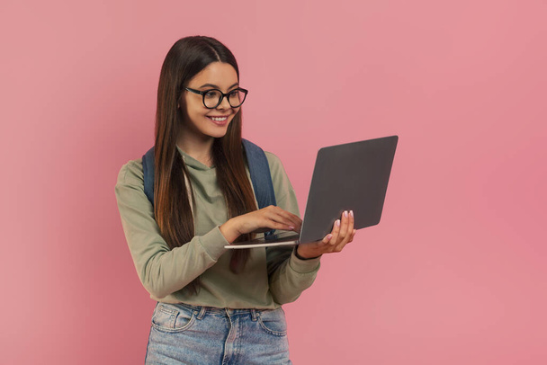 Smiling teenage girl wearing eyeglasses and backpack looking at laptop screen, cute female student symbolizing education and online learning, standing against minimalistic pink background, free space - 写真・画像