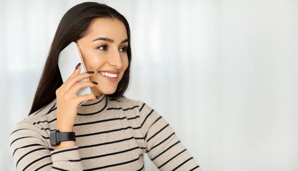A smiling young woman in a striped shirt engaging in a pleasant conversation on her smartphone, exuding confidence and positivity in a bright setting. Communication, gossip - Photo, Image