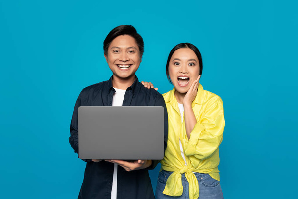 Surprised and happy millennial Asian couple with a laptop, the woman touching her face in excitement and the man smiling, indicating good news or success on a cyan background - Fotoğraf, Görsel