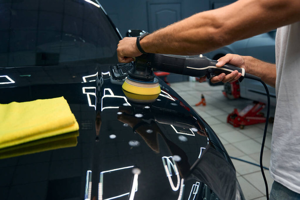 Man in car repair shop polishes the hood of a black car, he uses a grinder and a soft napkin - Fotó, kép
