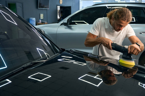 Long-haired young man polishes the body of a black car, he uses a special device - Foto, Imagen