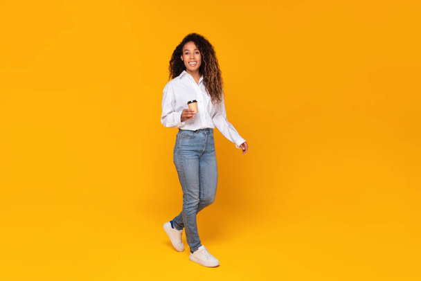 Full length shot of cheerful black lady holding paper coffee cup while walking and smiling to camera on yellow studio background, enjoying her break and tasty drink concept - Фото, зображення