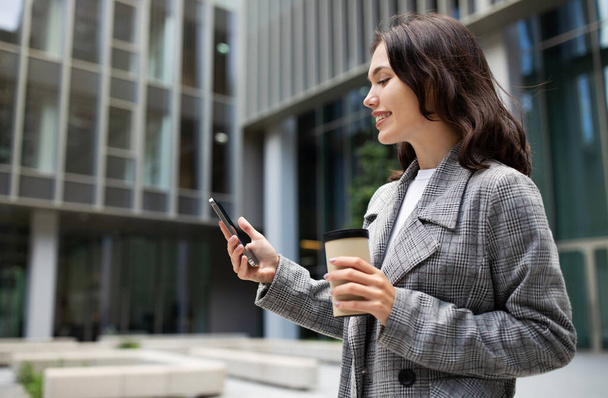 Business Application. Caucasian entrepreneur woman standing with coffee and phone, outside business center, messaging and scrolling social media app. Side view, copy space - Fotoğraf, Görsel