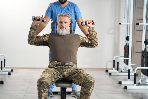 Bearded man is working out in the gym in a rehabilitation center, an experienced physiotherapist is working with him - Fotoğraf, Görsel