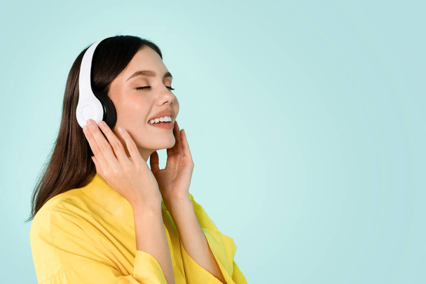 Cheerful millennial brunette woman in casual and wireless headphones enjoy listening music with closed eyes, isolated on blue studio background, copy space. Fun, relax, audio app, ad and offer, hobby - Foto, Bild