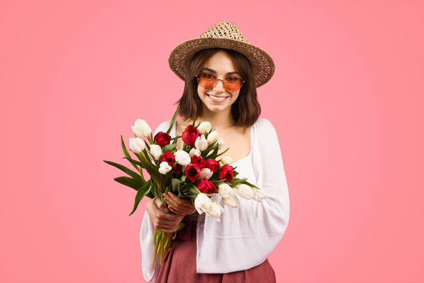 Radiant glad european young woman with a joyful smile, wearing a straw hat and round sunglasses, holding a bouquet of fresh red and white tulips against a soft pink backdrop - Фото, изображение