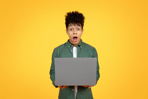 Online education, e-learning. Amazed young black guy using laptop, typing on computer keyboard, looking at camera and grimacing, copy space for advertisement, yellow studio background - Φωτογραφία, εικόνα