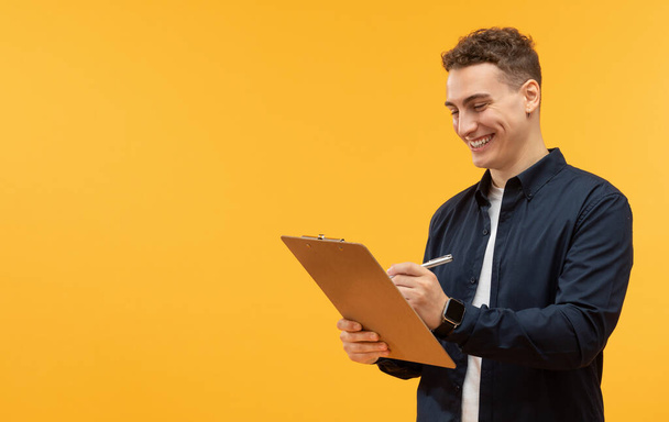Happy handsome young man taking notes in clipboard, yellow studio background, copy space for advertisement. Guy worker employee writing down on board, filling form - Фото, зображення