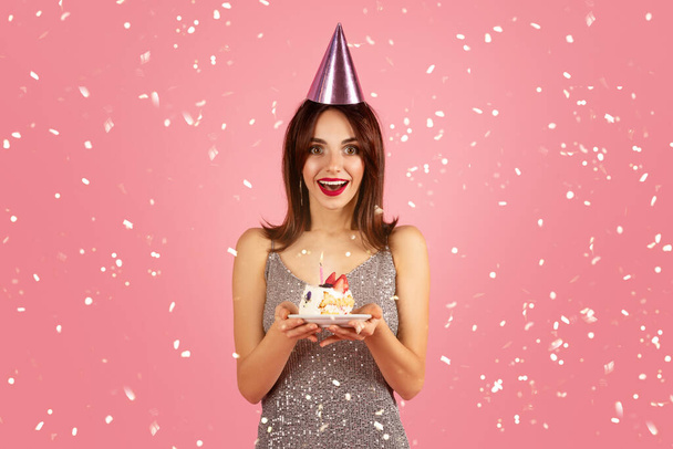 Excited woman with brown hair and red lipstick, wearing glittering silver dress and pink party hat, is delightedly hold birthday cake with one candle, with confetti falling on pink background - Фото, изображение