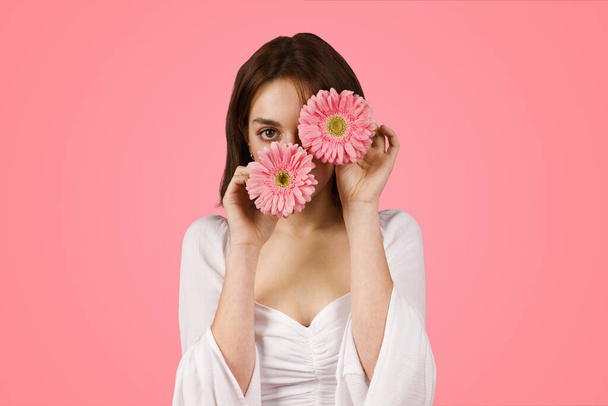 Mysterious and attractive caucasian young woman playfully holding two pink gerbera daisies over her eyes, creating a whimsical vibe against a striking pink studio backdrop - Photo, Image