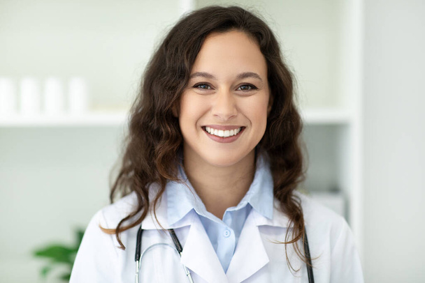 Closeup portrait of cheerful attractive brunette young european woman doctor wearing medical uniform white coat and stethoscope posing at clinic or hospital, smiling at camera - Фото, зображення