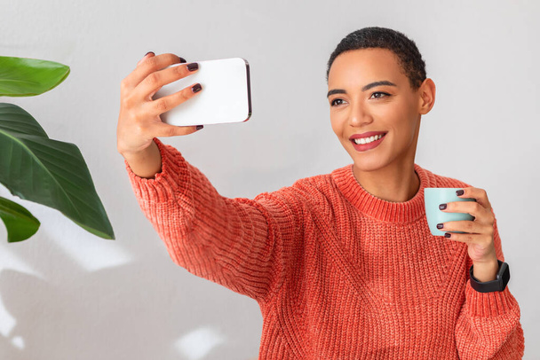 Radiant happy latin young woman taking a selfie on phone, holding a mug with a joyful smile, wearing an orange sweater, with a backdrop of lush green plants in a bright room - Фото, изображение