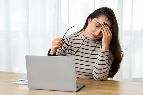 An exhausted young woman holding her eyeglasses feels a headache while sitting at her workspace with a laptop, indicating stress or overwork. Lady suffer from pain, health problems - Photo, image