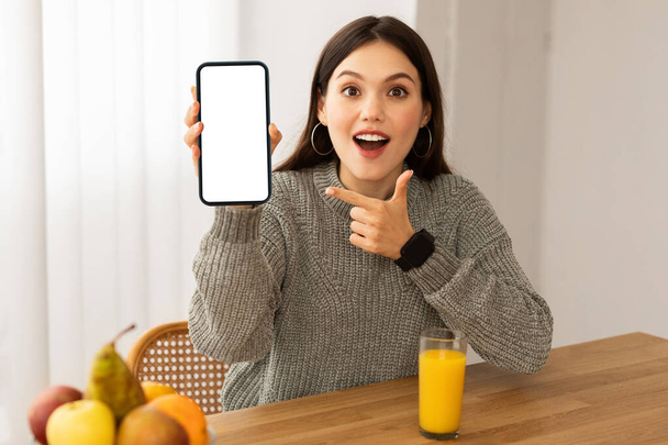 Amazed beautiful young woman showing nice mobile app for weight loss, sitting at table with fresh organic fruits and orange juice, holding smartphone with white blank screen, mockup, copy space - Фото, зображення