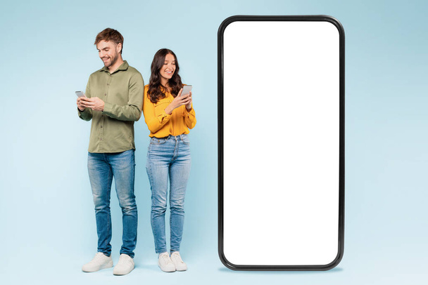 Man and woman using smartphones beside blank mockup screen, perfect for presenting apps or advertisements, with friendly, tech-savvy vibe against blue background - Photo, Image