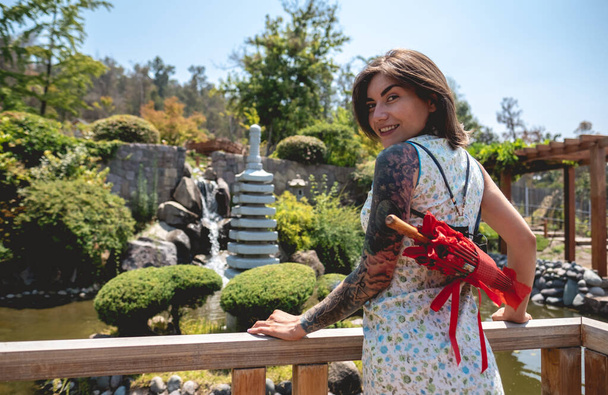 Lenyűgöző szépség és japán stílus: barna lány tetoválásokkal, ázsiai ruha, és piros papír esernyő, élvezet egy napsütéses napon kék ég egy japán kertben - Fotó, kép