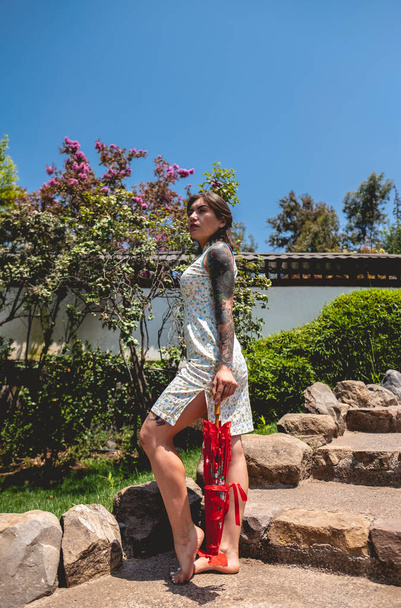 Impresionante belleza y estilo japonés: chica morena con tatuajes, vestido asiático y paraguas de papel rojo, deleitándose en un día soleado con cielo azul en un jardín japonés - Foto, imagen