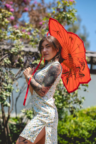 Upea kauneus ja japanilainen tyyli: brunette tyttö tatuoinnit, aasialainen mekko, ja punainen paperi sateenvarjo, iloksi aurinkoisena päivänä sininen taivas japanilainen puutarha - Valokuva, kuva