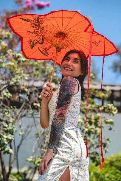 Oszałamiająca uroda i japoński styl: brunetka z tatuażami, azjatycka sukienka i czerwony papierowy parasol, zachwycający w słoneczny dzień błękitnym niebem w japońskim ogrodzie - Zdjęcie, obraz