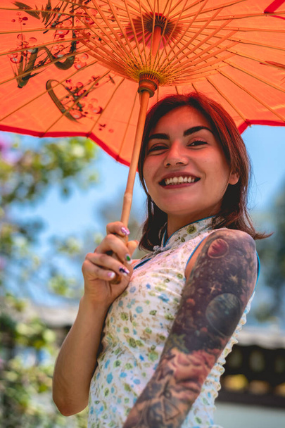 Lenyűgöző szépség és japán stílus: barna lány tetoválásokkal, ázsiai ruha, és piros papír esernyő, élvezet egy napsütéses napon kék ég egy japán kertben - Fotó, kép