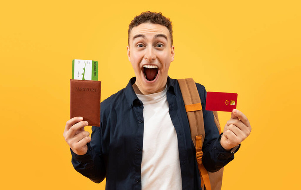 Happy excited guy student showing red plastic bank credit card and passport with flight tickets, carrying backpack, going vacation, travelling abroad, yellow studio background - Photo, Image
