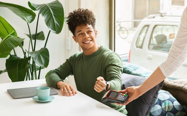 Positive stylish african american young man using smart watch while making contactless payment in cafe. Cheerful black guy freelancer working at coffeeshop, paying for service with banking mobile app - Fotoğraf, Görsel