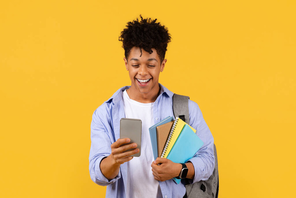 Delighted black male student engaging with smartphone while holding notebooks, reflecting blend of traditional and digital education on yellow background - Foto, afbeelding