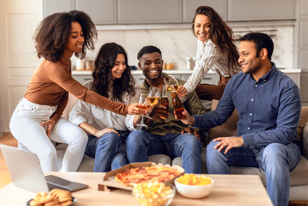 Diverse group of students clinking with bottles and glasses of beer, sharing stories and laughs in a modern living room setting, enjoying party gathering with pizza, sitting on sofa indoors - Photo, Image