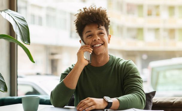 Smiling handsome african american guy sitting at cozy cafe next to window, talking on phone, drinking coffee, looking at copy space. Black millennial man working at coffee shop. Communication - Foto, Imagem