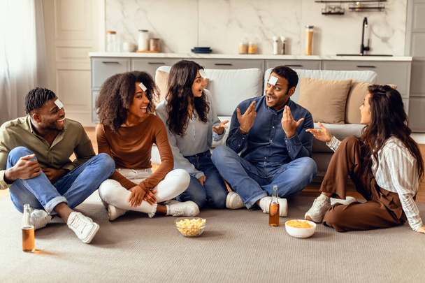 Multiracial group of young friends playing Guess Who game in the living room, enjoying joyful weekend gathering, sitting in circle on floor, having papers on foreheads. Weekend leisure - Fotoğraf, Görsel