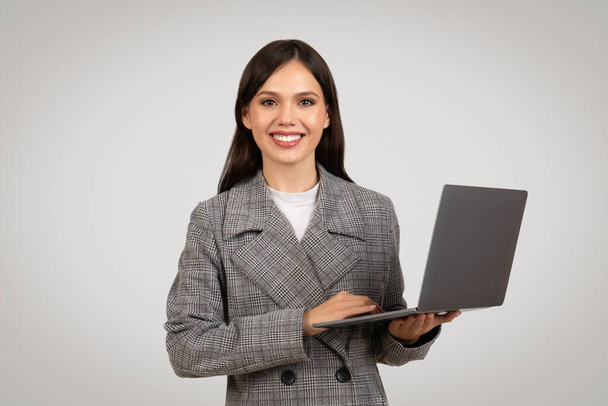 Confident young businesswoman in checkered suit holding an open laptop with friendly smile, representing modern professionalism and tech proficiency - 写真・画像