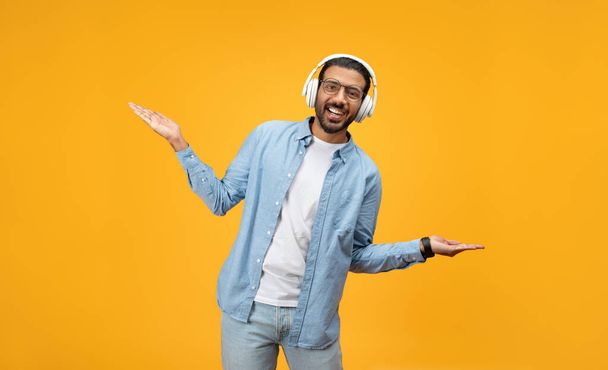An exuberant man in a denim shirt wears white headphones and gestures with his hands, showing excitement or dancing to music, against a vibrant orange background - Zdjęcie, obraz