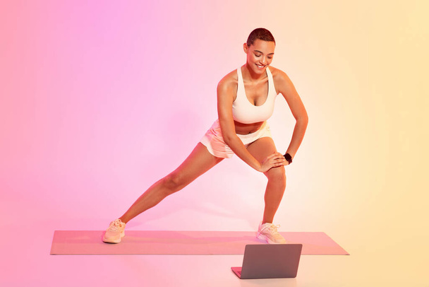 A focused woman with a shaved head stretches on a yoga mat in a side lunge position, looking at a laptop screen, wearing a white sports bra and shorts against a colorful background - Foto, Bild