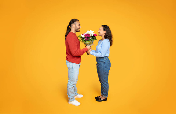 A smiling man in a red sweater gives a lush bouquet to a woman in blue, both radiating happiness against a cheerful yellow background, celebrating lovers day, full length - Fotó, kép
