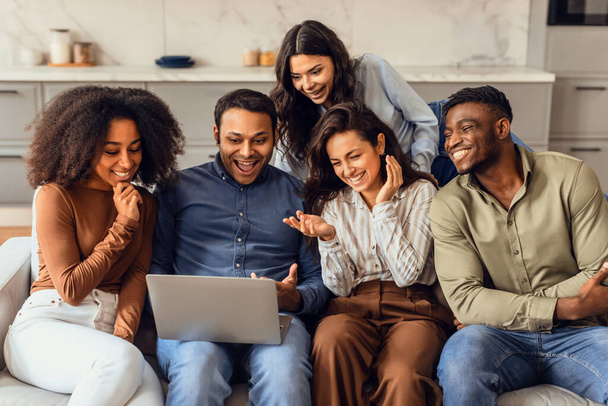 Group of young multiethnic friends watching funny comedy movie on laptop gathered in modern living room indoor. Fellow students enjoying weekend browsing and watch videos online - Foto, Imagem
