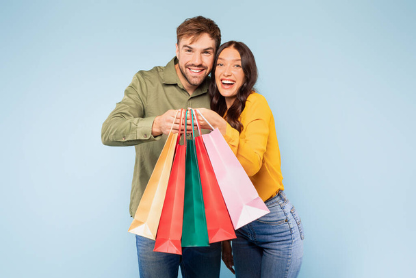 Radiant young couple holding collection of colorful shopping bags, their faces beaming with the thrill of successful shopping day, set against bright blue background - Photo, image