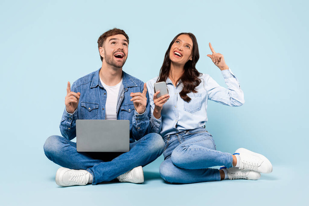 Enthusiastic young couple seated with laptop and smartphone, looking upwards with bright ideas, symbolizing moment of inspiration and creativity, against blue backdrop - Foto, imagen