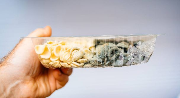 A male hand is holding a package of perished pasta, where half of the contents are still good, while the other half is covered with mold - Foto, Imagen