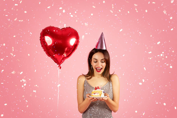 Delighted woman in a sequin dress with a party hat looking at a birthday cake with excitement, with a red heart-shaped balloon and confetti in the background. Holiday celebration event - Foto, imagen