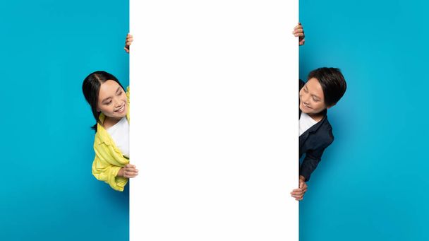 Asian millennial man and woman playfully peeking around a large white vertical banner, with joyful expressions, perfect for advertising space on a bright turquoise background - Fotoğraf, Görsel