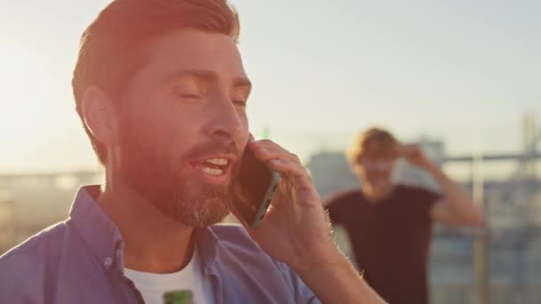 Hombre del partido llamando a smartphone en el primer plano de la azotea de verano. Sonriente tipo barbudo hablando teléfono celular sosteniendo botella de cerveza a la luz del sol de la noche. Feliz hipster relajado tener conversación telefónica en amigos pasar el rato - Imágenes, Vídeo