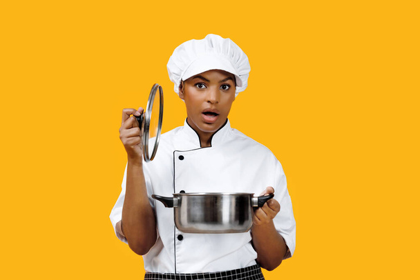 Surprised black female chef lifting the lid off pot and looking at camera, african american cook woman capturing her unexpected reaction to the dish, standing against bright yellow background - 写真・画像